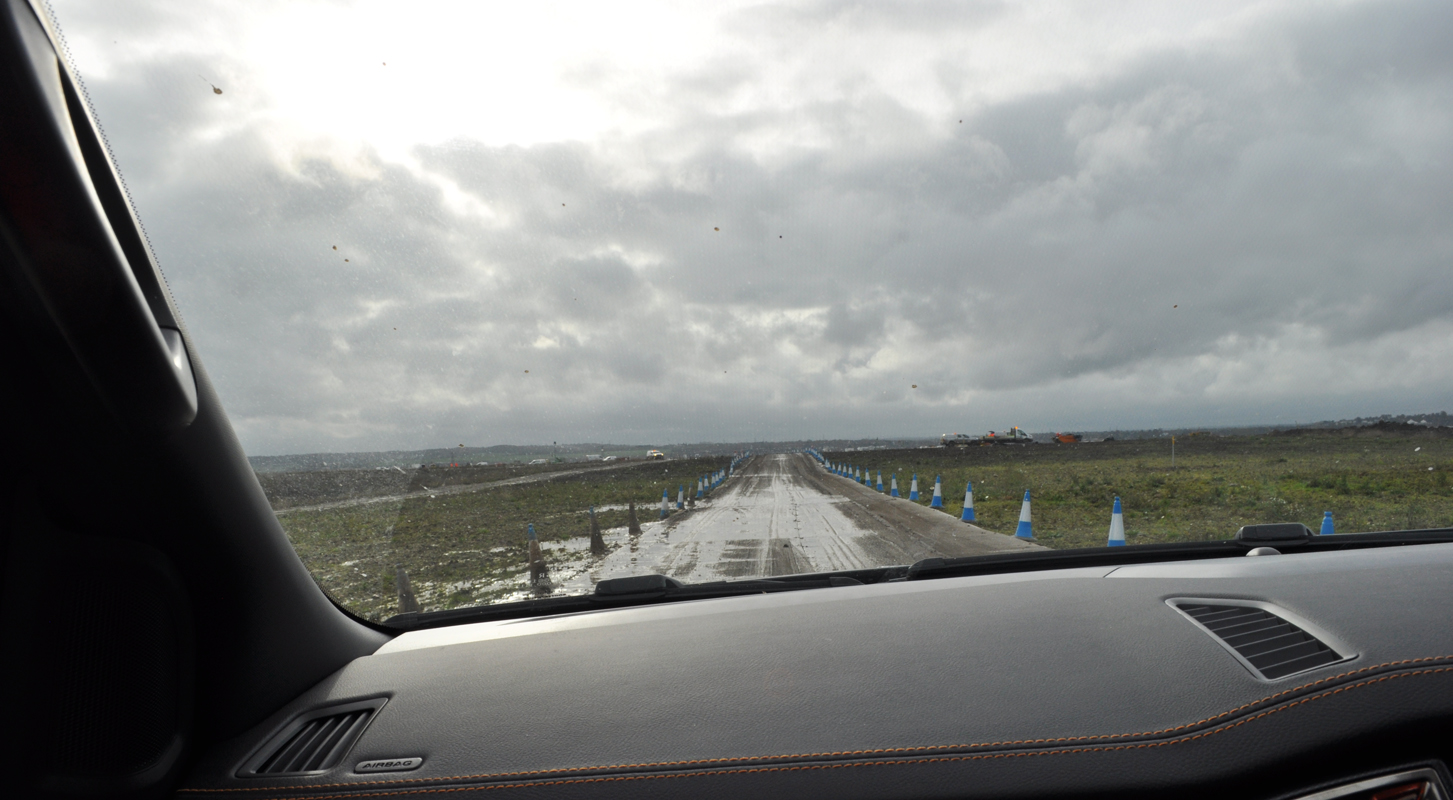 Lower Thames Crossing Ground Investigation Site Visit - A view of a very wet and muddy field with plastic track road laid on top, covered in puddles, edged with blue and white bollards. Photo taken from inside a vehicle looking out of the front window